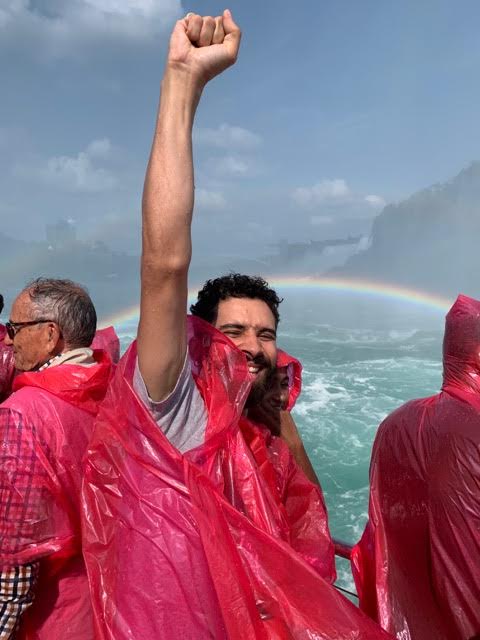 Malik, in a bright pink rain poncho, raises a hand above his head in front of a rainbow.