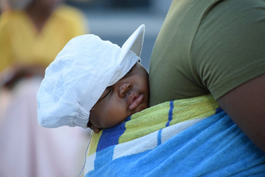 A young child with a white hat sleeps while cradled on their parent.