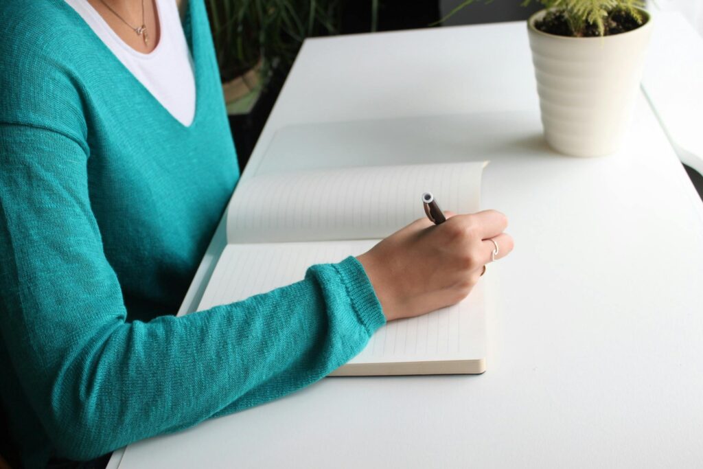 A picture of a faceless person in a blue sweater sitting at a desk, preparing to write in a notebook.