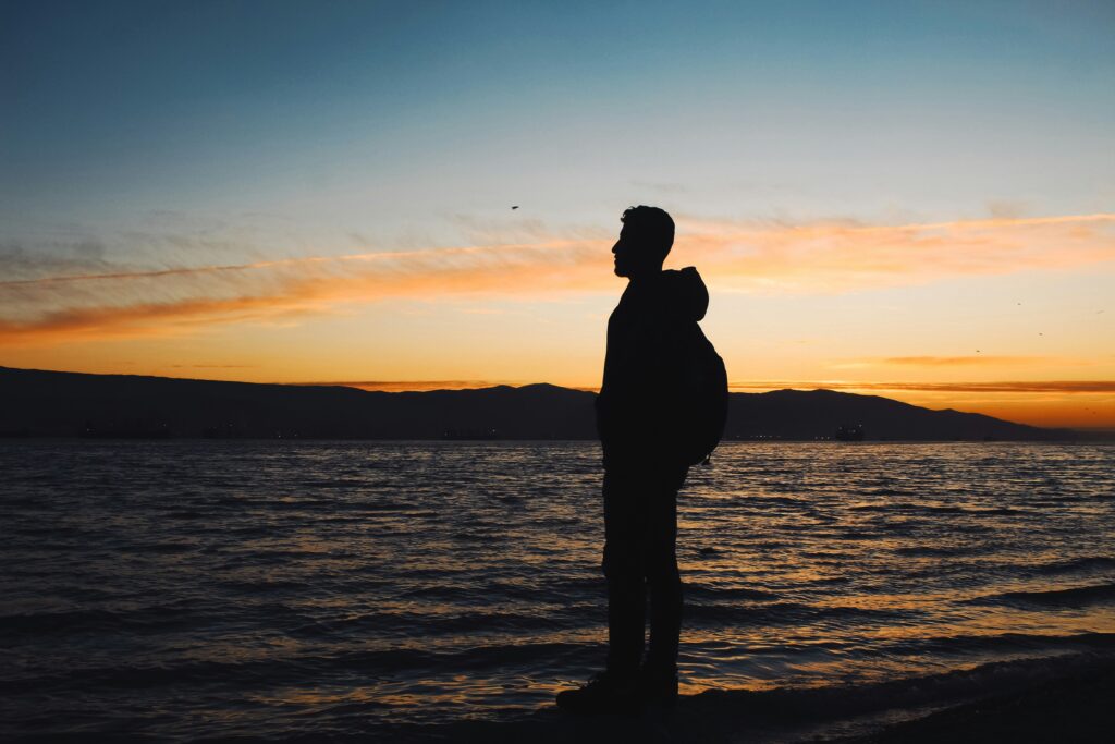Profile of a man standing on a beach, with a sunset behind him. 