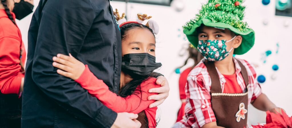 A young man in his twenties smiles as he receives a warm hug from a young girl wearing a festive holiday headpiece and face mask. A boy wearing a hat fashioned like a Christmas tree stands nearby looking at the camera (also masked).