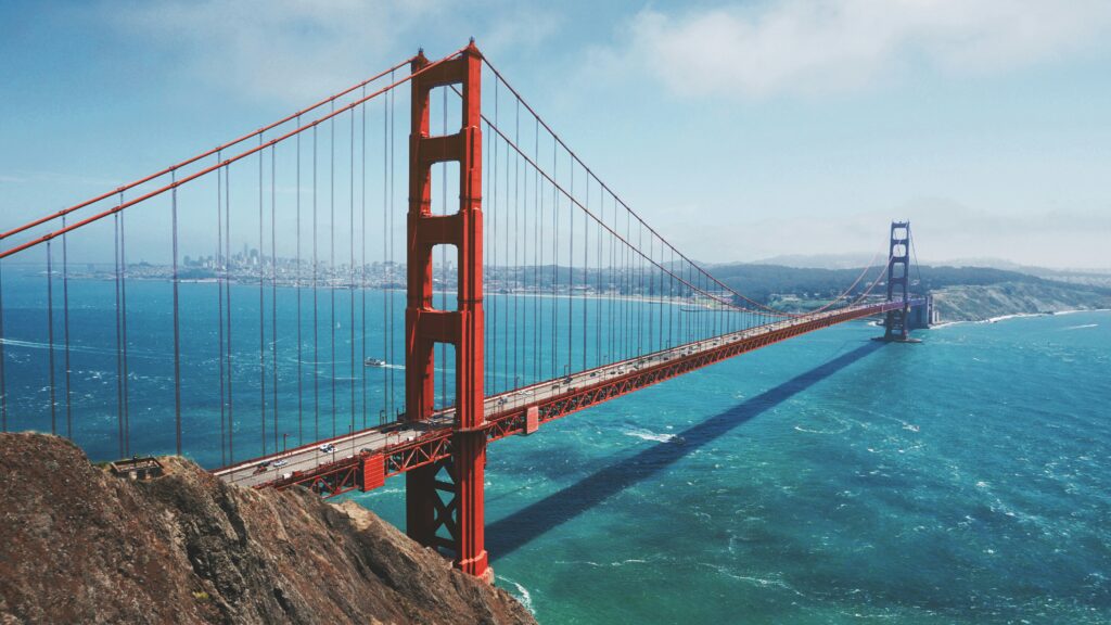 A wide shot of The Golden Gate Bridge in San Francisco, California. The weather is slightly cloudy and the water beneath the bridge is a mix of different blues, due to the current. At the left of the bridge are some cliffs. In the distance, the city skyline is visible. 
