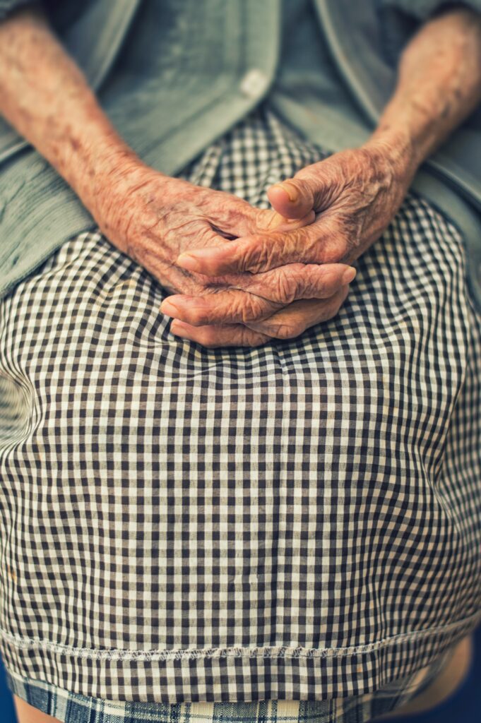 Image shows the clasped hands in the lap of an old woman, resting on her black and white checkered skirt.