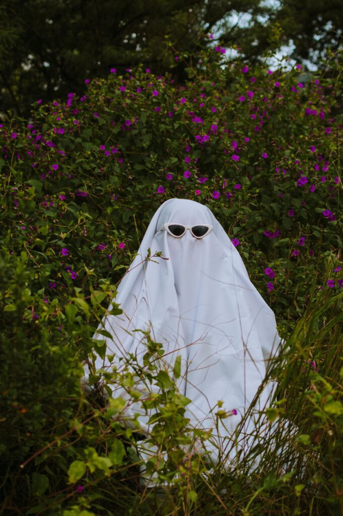 A ghost wearing sunglasses hiding in a field of shrubs and greenery with purple flowers.