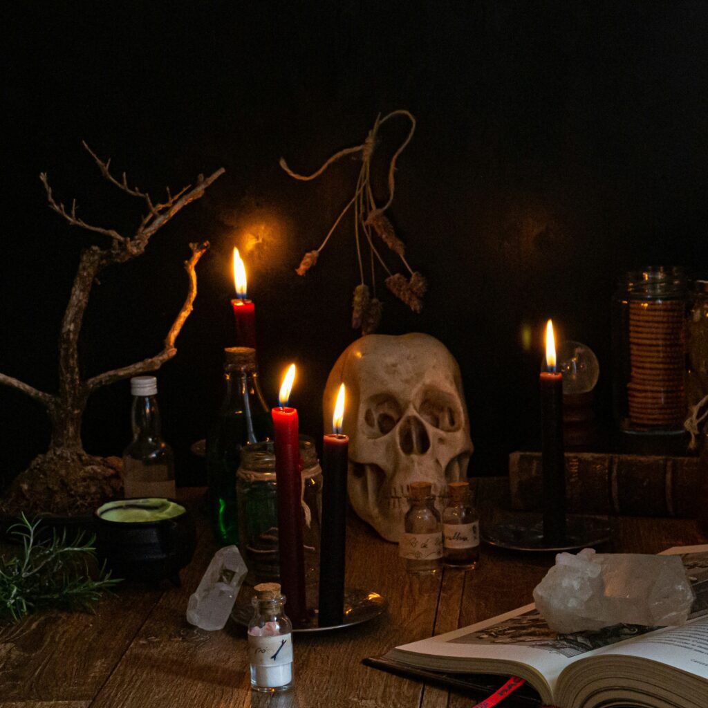 A skull, red candles, a spell book, and potions/vials sit on an old table – a scene fit for a witch and other figures of the night!