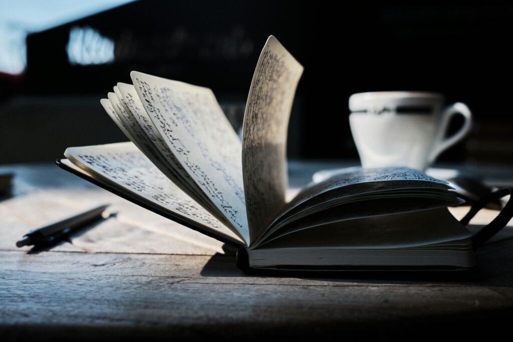 A journal splayed open. To the left of the journal is a pen. Behind the journal is a teacup.