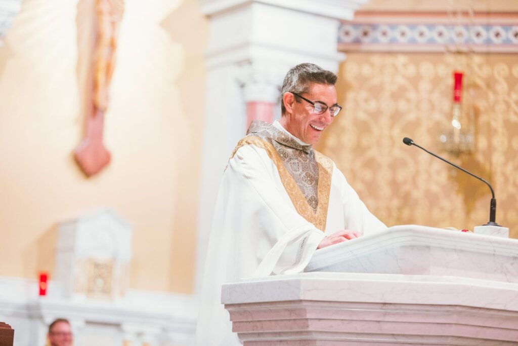 Image of Father Game Hammerschmidt of St. Isidore’s Catholic Church in Manhattan, Kansas. He is at the altar, smiling, and looking downward.