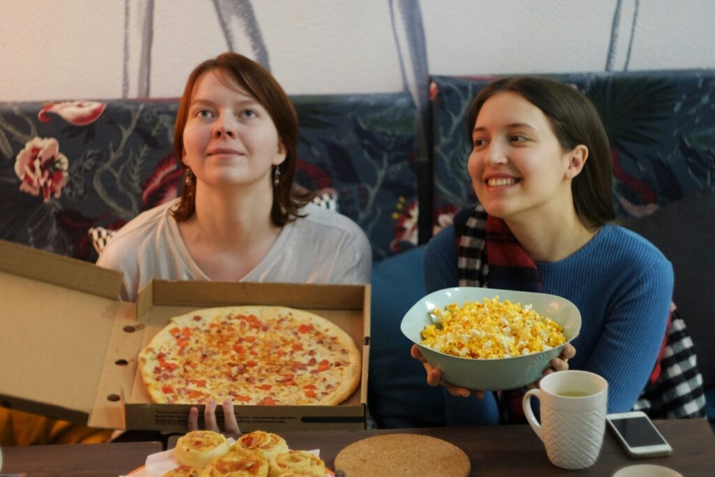Image of two young woman sitting on a couch. One is holding a pizza in a box, while the other is holding a bowl of popcorn.