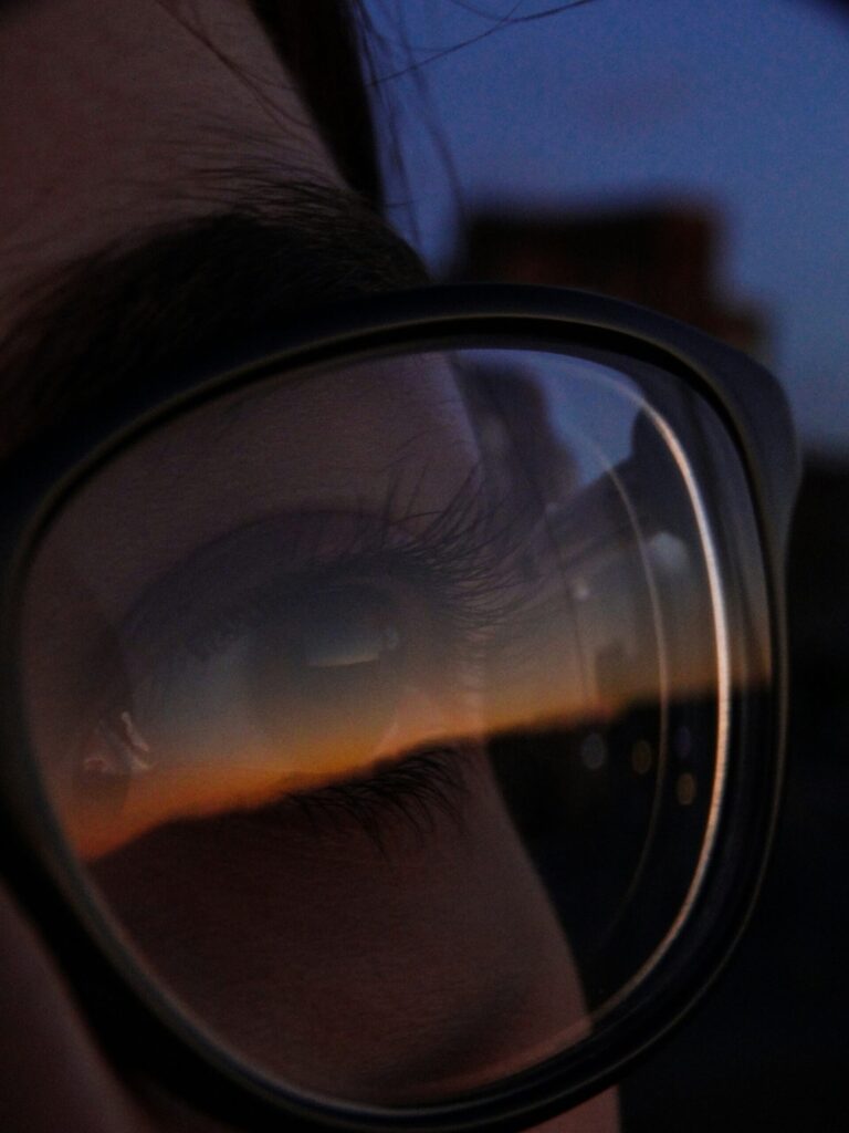 Image of a woman’s eye, slightly hidden behind a large pair of sunglasses. The sunset is reflected on her glasses lens.