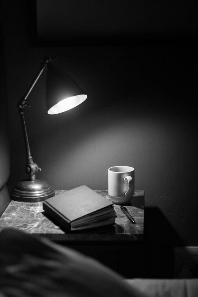 Image of a sidetable with a lamp, journal, pen, and coffee mug on it. The image is in greyscale.