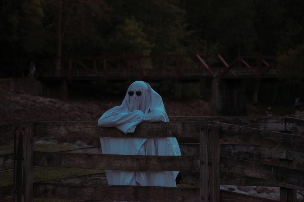 A white sheet ghost with sunglasses leans, with crossed arms, on a wooden fence staring off into the distance.