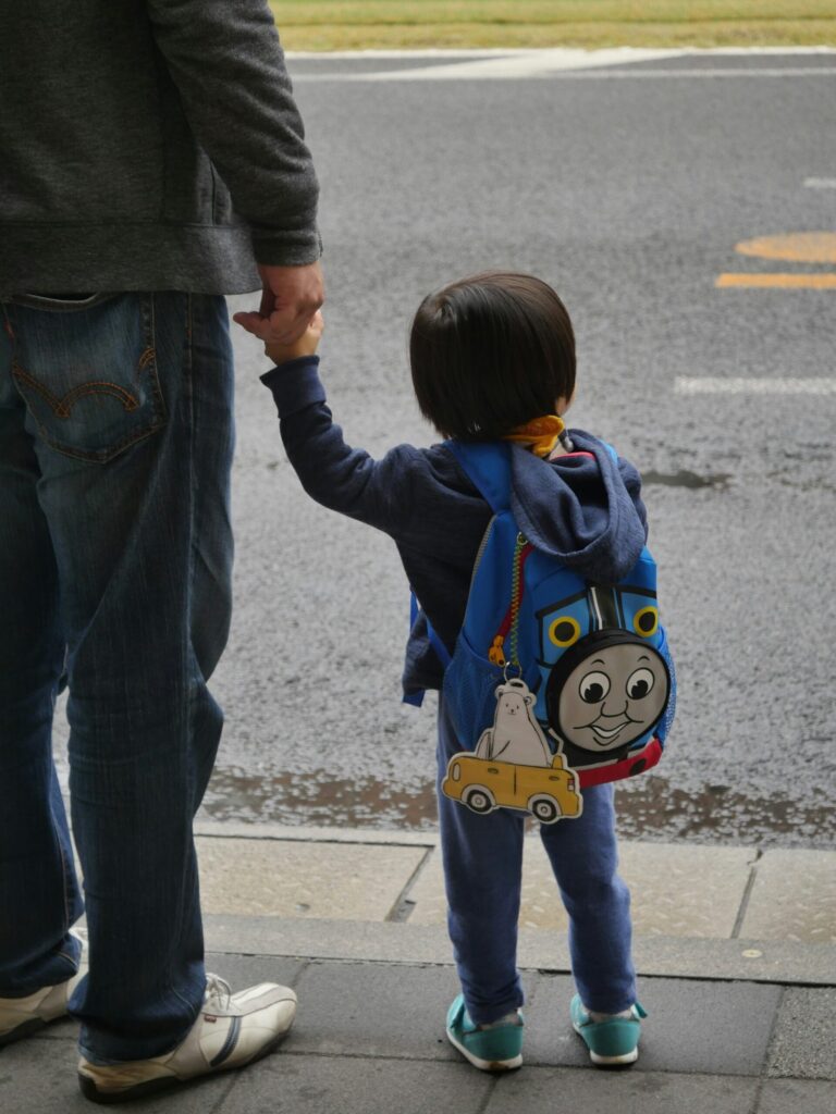 Photographed from behind, a man holds the hand of a kindergartener on the side of a street. The child wears a Thomas the Tank Engine backpack, and looks out across the street.
