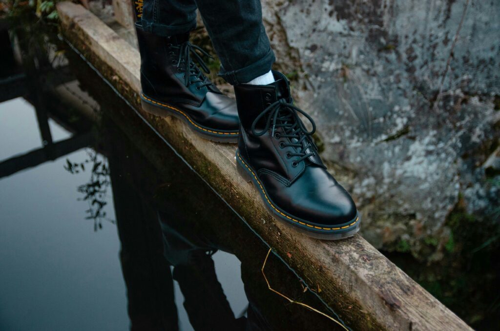 A person wearing black Doc Martens while walking along a wooden beam over water.