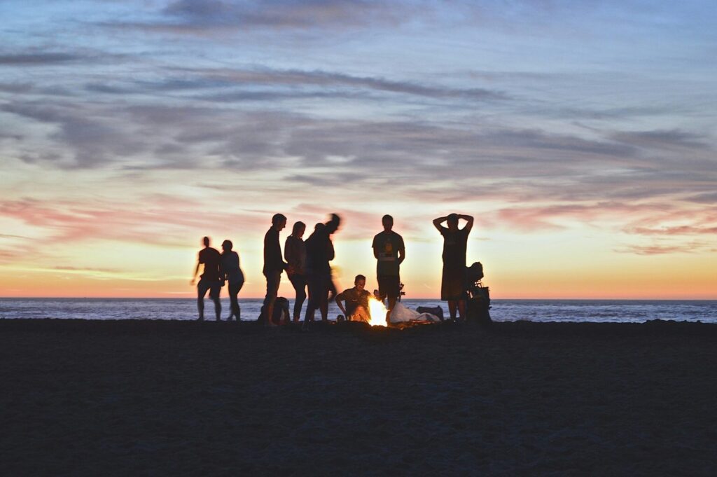 Image of a group of friends hanging out on a beach. In the background, the sun is setting.