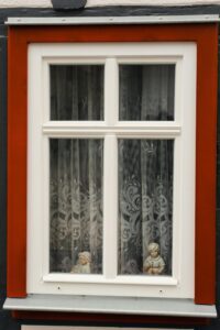 A red wall with a white window housing a lace curtain and two child figurines staring out onto the world.