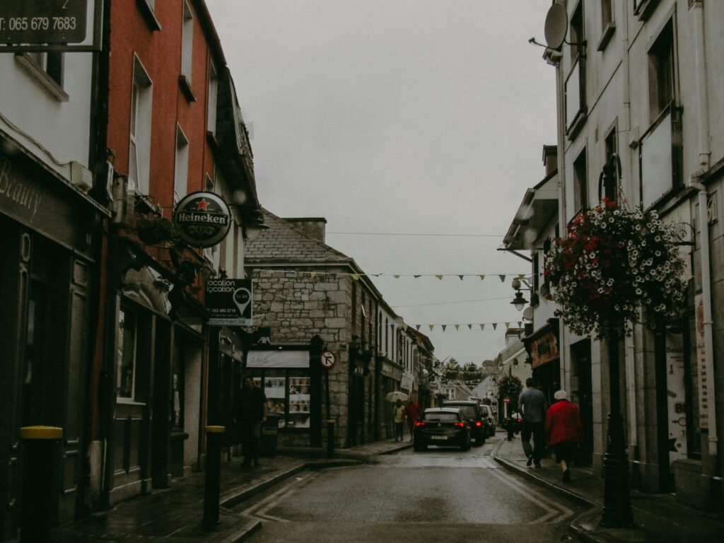 Aged town buildings line a narrow cobblestone street on a cloudy, chilly day, with few people and vehicles in sight.