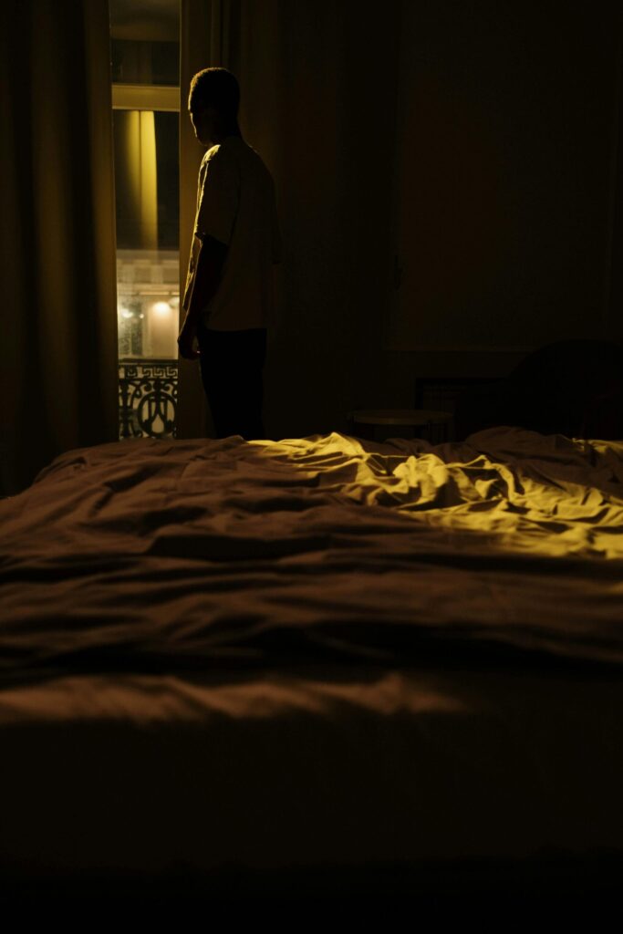 A person stands in the dark of their bedroom while street lights cast a yellow glow into the room.