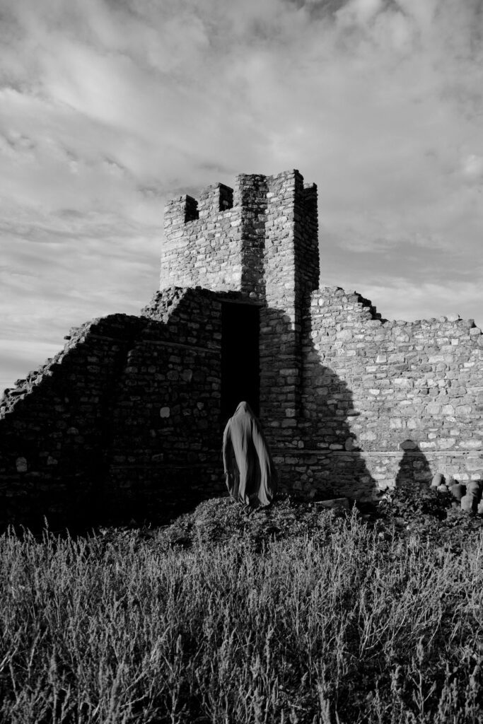 A grayscale cloaked figure looms in the shadows of a crumbling castle wall.
