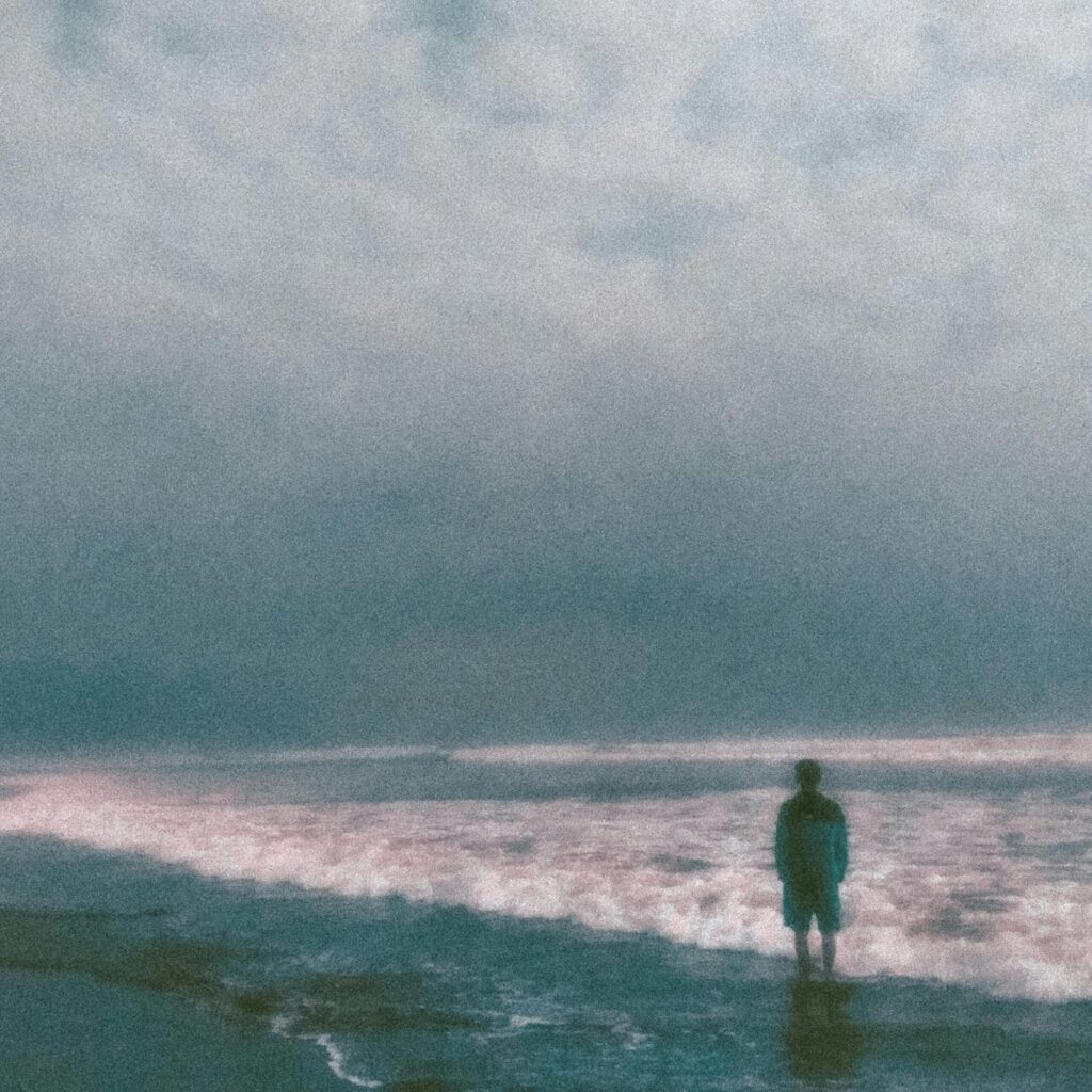 A photo of a lone figure standing in a wave on a cloudy beach.