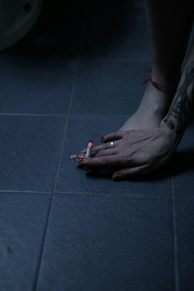 A woman’s hand rests on her foot and the cold tile floor with a lit cigarette between her fingers.