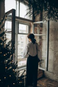A woman stands, staring out of a large window, while evergreen boughs dressed in lights frame the scene.