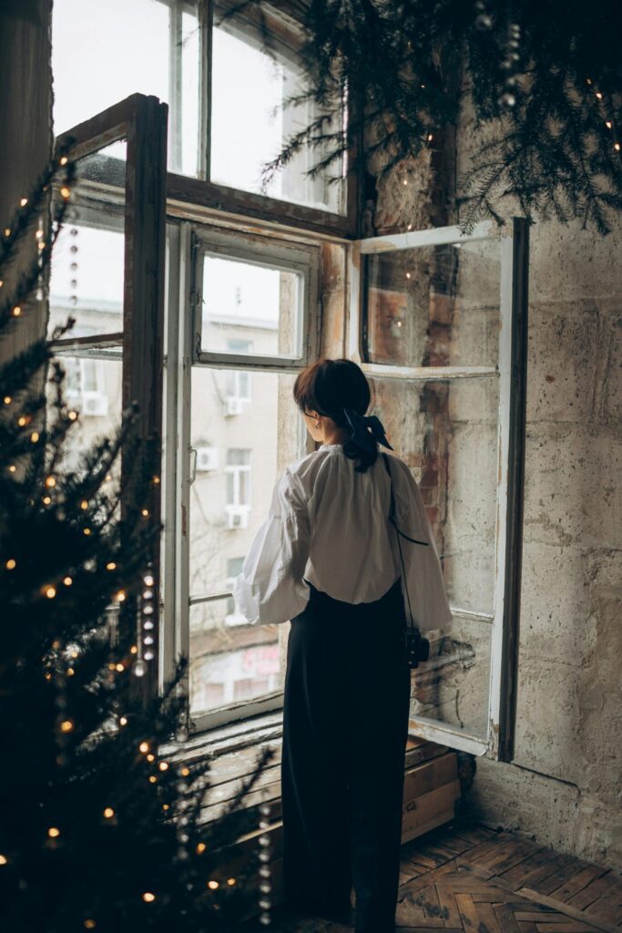 A woman stands, staring out of a large window, while evergreen boughs dressed in lights frame the scene.