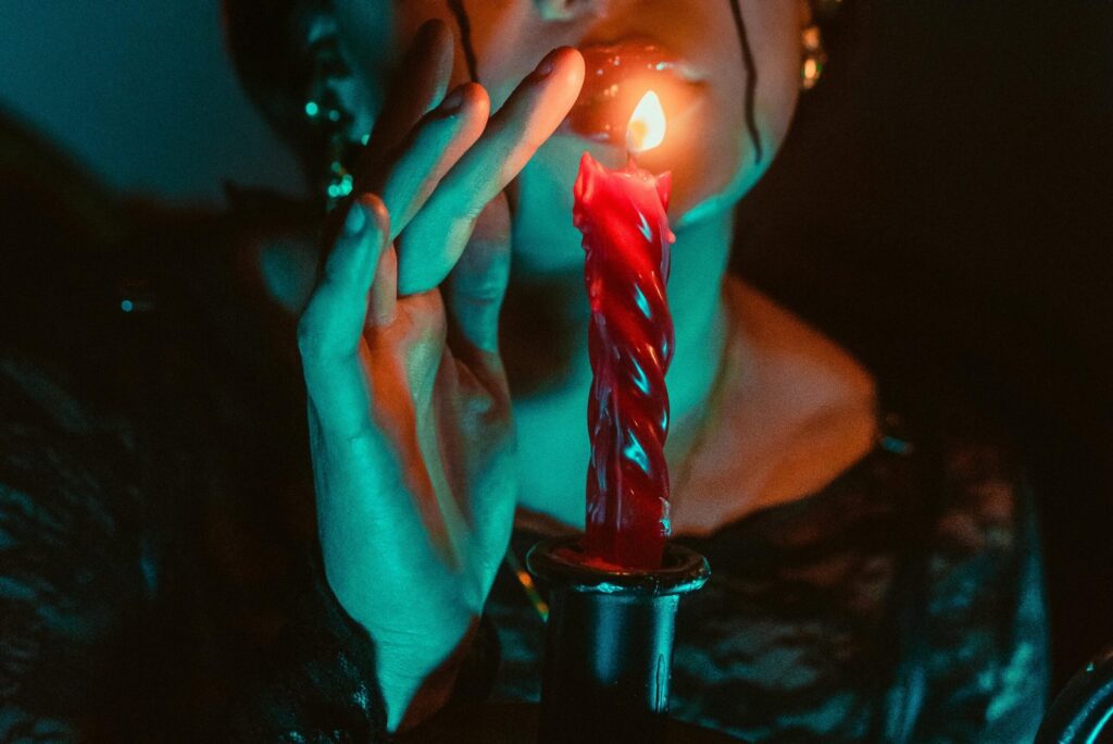 A woman, dressed in black, holds her finger over a blood-red candle that drips slowly. The entire scene is lit in a ghoulish green-blue light.