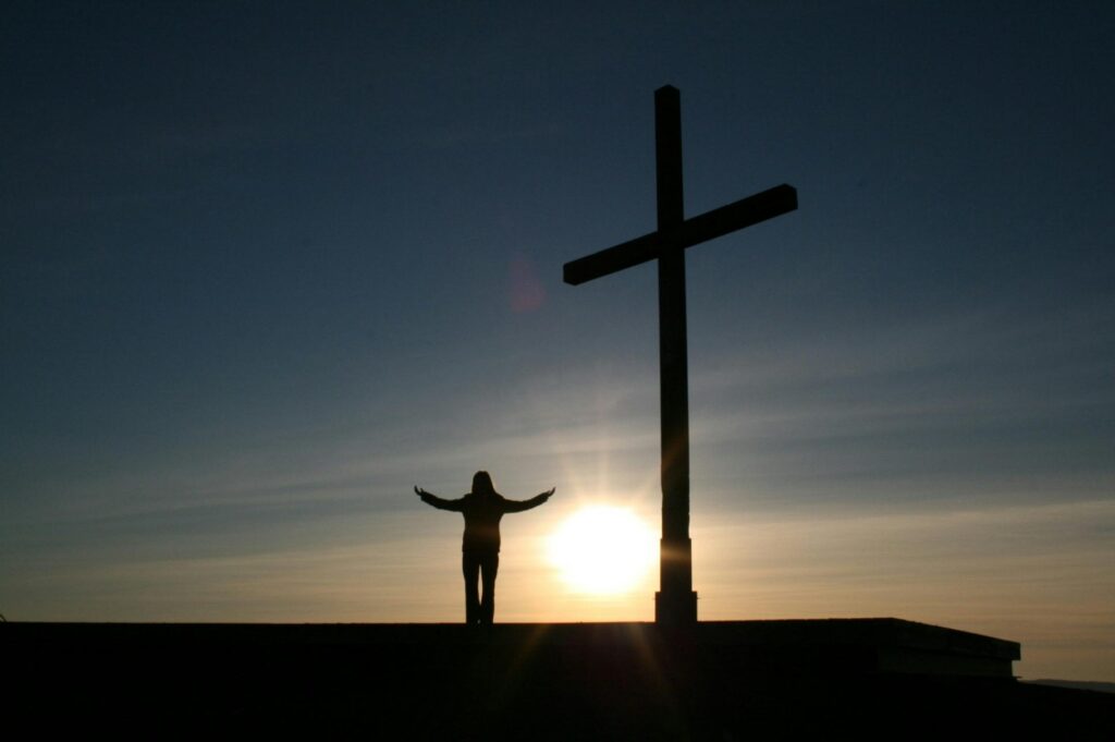 The silhouette of a person standing beside a gigantic cross during sunset.