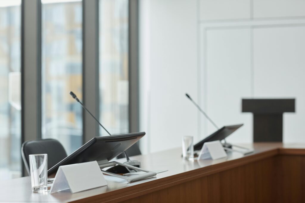An image of two monitors and long, skinny microphones atop a conference table with a podium in the background.