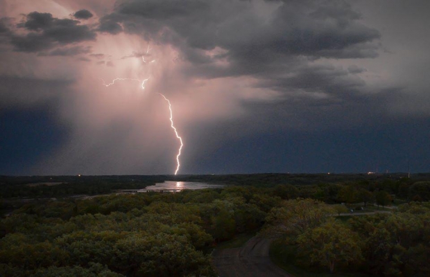 Lightning strikes beyond a river and a green landscape at dusk.