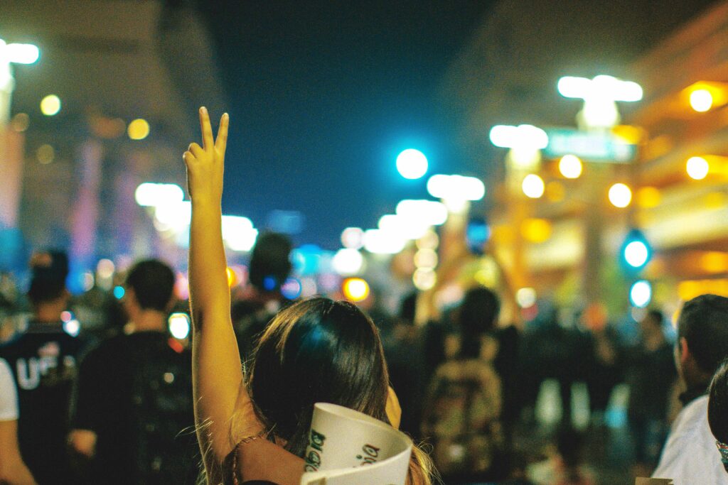 Image of a woman at a protest. She has her back turned to the camera, and she’s raising her left hand, which is shaped as the peace sign.