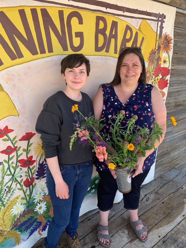 Two people standing next to one another, smiling. One woman is holding a vase of flowers. 