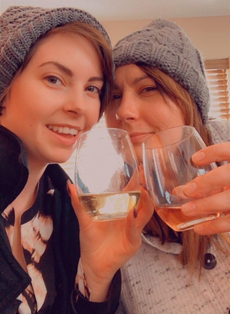 Two people cheersing two wine glasses together, wearing matching gray cable-knit beanies. 