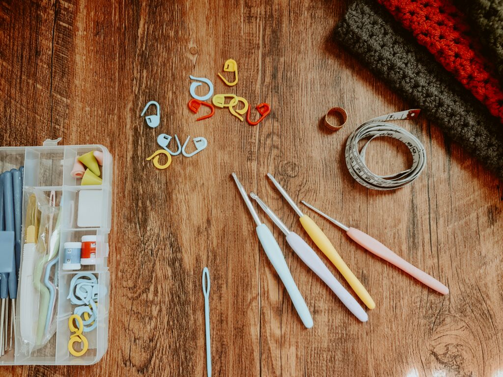 A tabletop covered with various notions associated with crocheting: stitch markers, a tape measure, crochet hooks, and a tapestry needle among them.