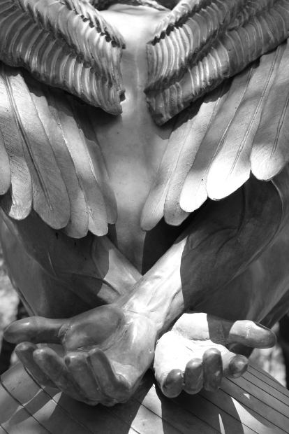 A black and white photo of a sculpture of an angel. The focus of the photos are the angel’s wings above two arms crossed over one another at the wrist, palms open.