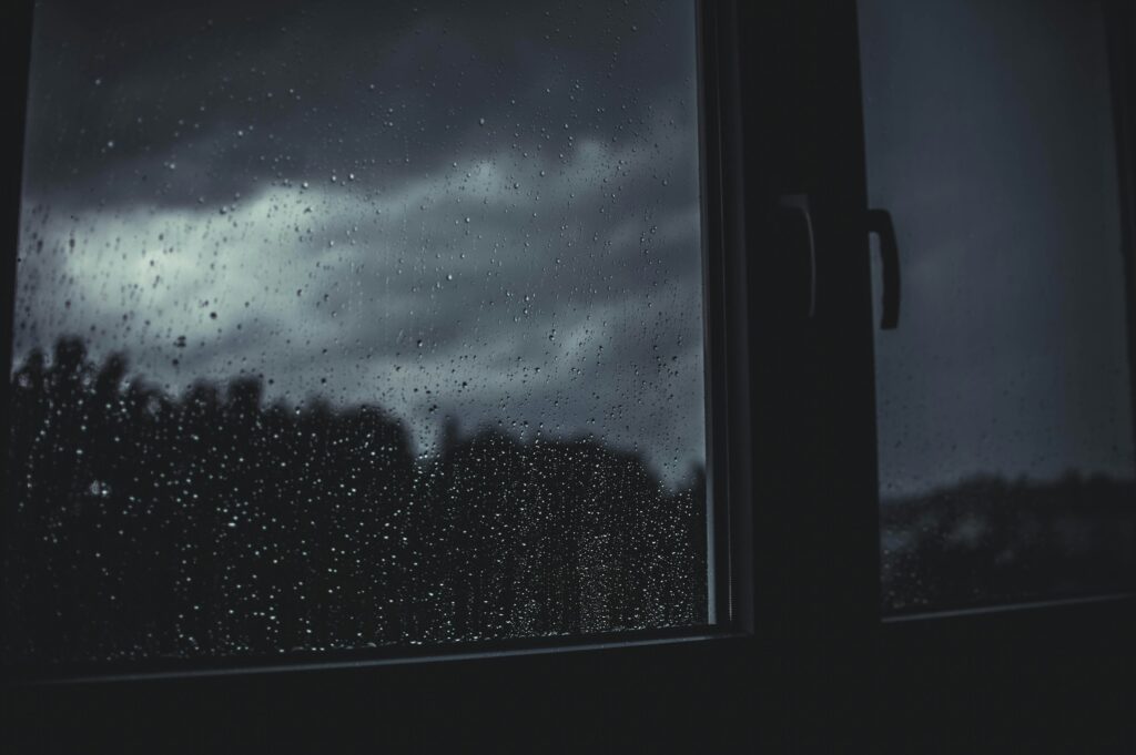 Image of a window with raindrops on the glass. Outside the window are stormy, grey skies. The entire photo is dark and grey.