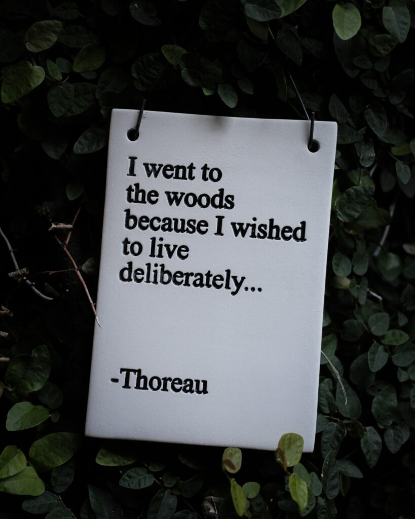 A white garden sign, among green leaves, has a famous Thoreau quote written in black text that reads, “I went to the woods because I wished to live deliberately…”