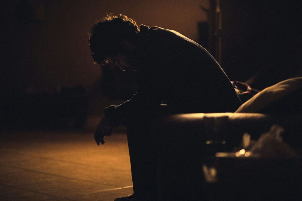 image of a person sitting down, hunched over. The image is dark with little light. Shadows cover most of the person’s body.