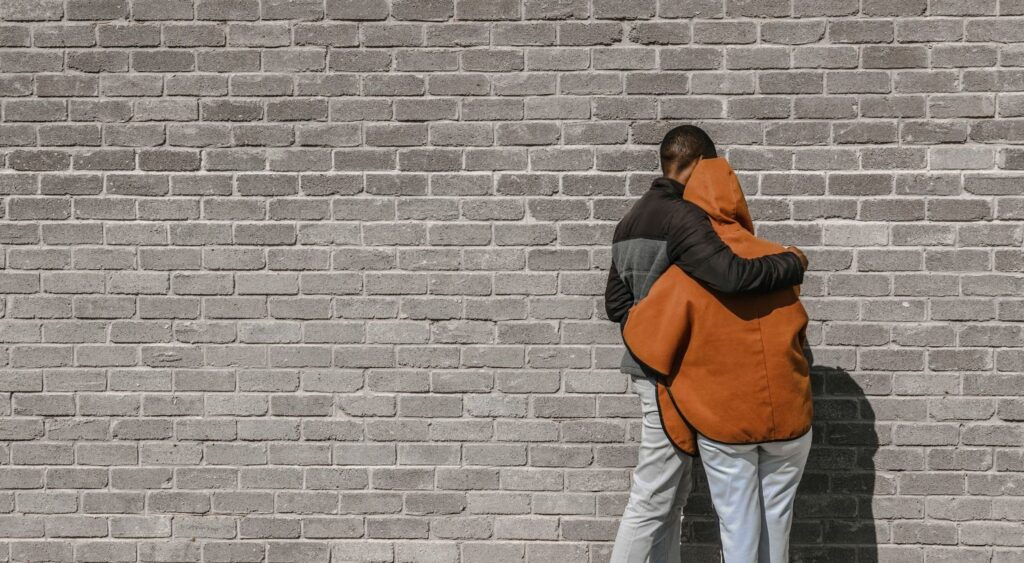  Image of two people hugging each other. Their backs are turned to the camera. They’re facing a brick wall.