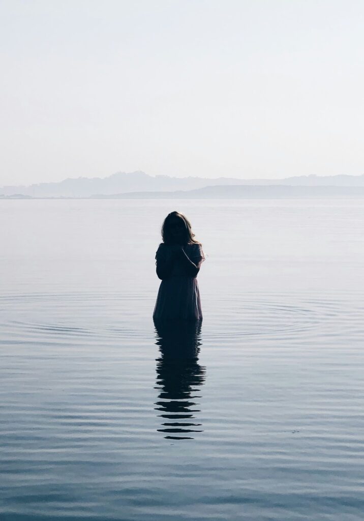 Image of a woman in a dress standing in a lake with her head in her hands. Shadows obscure her entire front, including her face.
