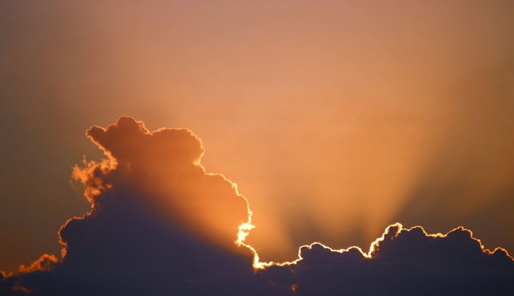 Image of grey clouds with the sun lighting up the edges of each cloud. The edges are a bright, coppery yellow and orange, contrasting the dark color of the rest of the clouds.