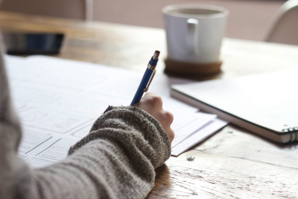Image of a person writing with a pen on paper. They’re seated at a table, and in the background sits a mug and a notebook.