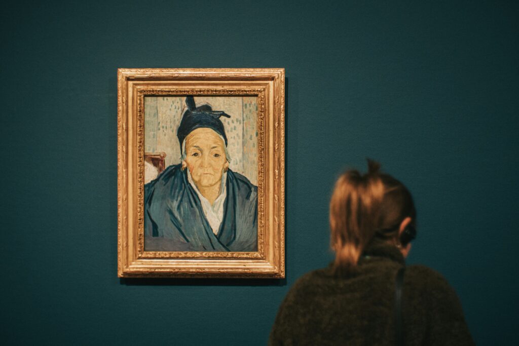 Image of a young woman looking at a painting that features an older woman. The young woman’s back is turned to the camera.