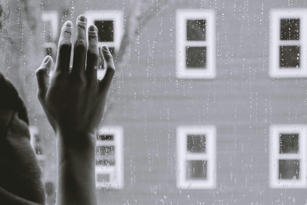 Image of a person putting their hand against a rainy window. Outside the window is another building with several windows.