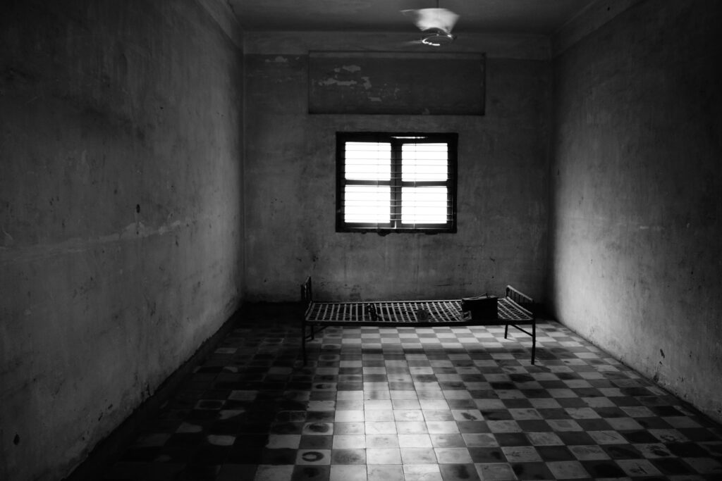 Image of an empty prison cell. There’s a metal cot in the middle of the cell without a mattress. Behind the metal cot, on the wall, is a single, four-pane window. The entire photo is black and white.