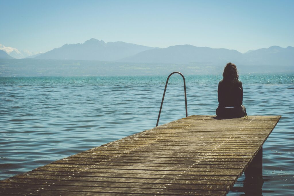 Image of a woman sitting at the end of a boat dock, staring at the water. In the background loom mountains.