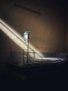 An empty hospital bed sits against a wall, IV bag hanging above. A sliver of sunlight illuminates the bed as the rest of the background is steeped in shadow.