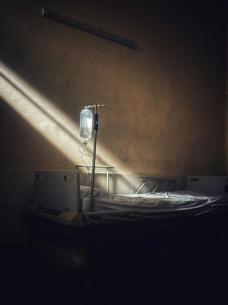 An empty hospital bed sits against a wall, IV bag hanging above. A sliver of sunlight illuminates the bed as the rest of the background is steeped in shadow.