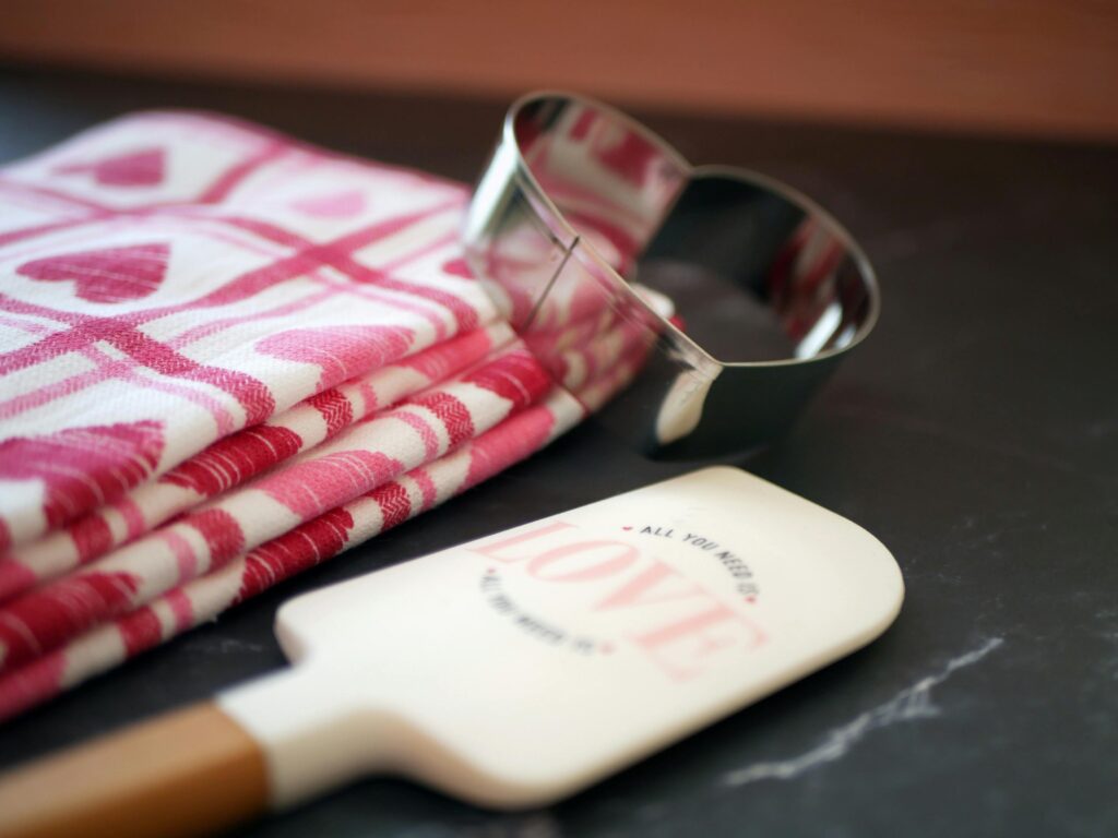 A heart-shaped cookie cutter, a hand towel printed with hearts, and a kitchen spatula that reads, “All you need is love.”