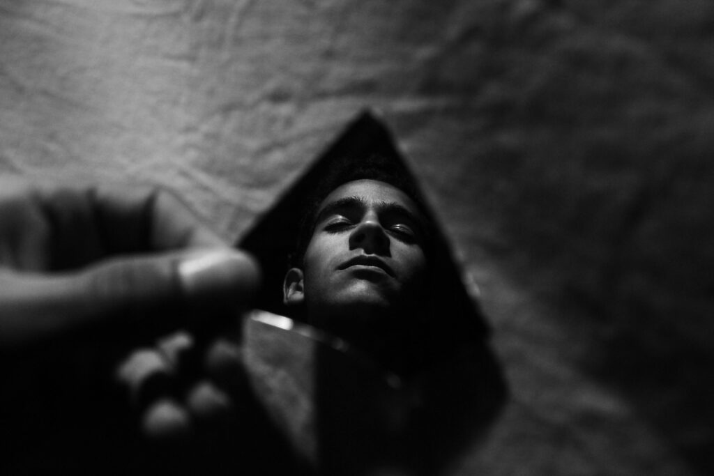 Image of a man holding a mirror shard in his hand. He’s staring at his reflection in the shard. The image is in greyscale.